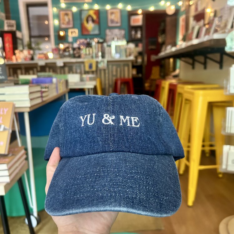 A hand holds the Yu & Me Hat by Yu & Me Books—a blue denim cap with “YU & ME” embroidery, shown in a cozy bookstore with yellow stools, bookshelves, and string lights—perfect for fans of unique hats.