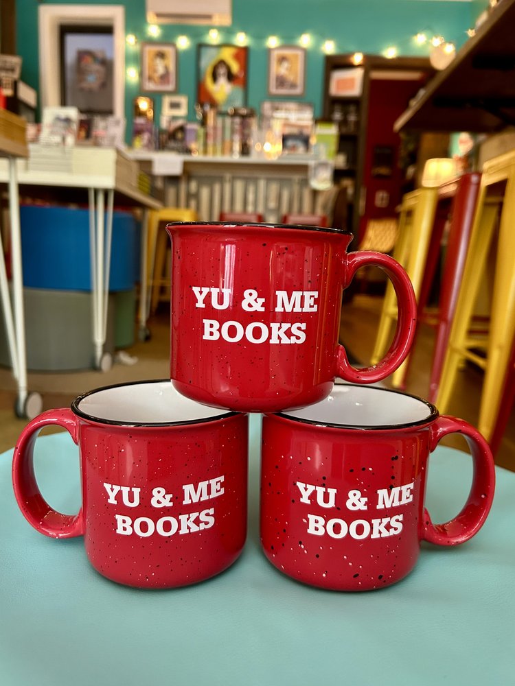 Three red Yu & Me Mugs by Yu & Me Books, each with white speckles and the bookstores name, are stacked in a triangle on a teal table—ready to hold your favorite warm drink in a cozy bookstore setting filled with books and string lights.