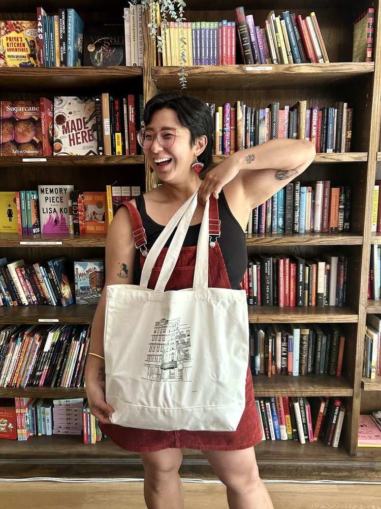 A smiling person in red overalls and glasses stands before colorful bookshelves, proudly displaying the Yu & Me Large Tote Bag by Yu & Me Books, featuring a Sammy Yuen building illustration and a vibrant book haul in the background.