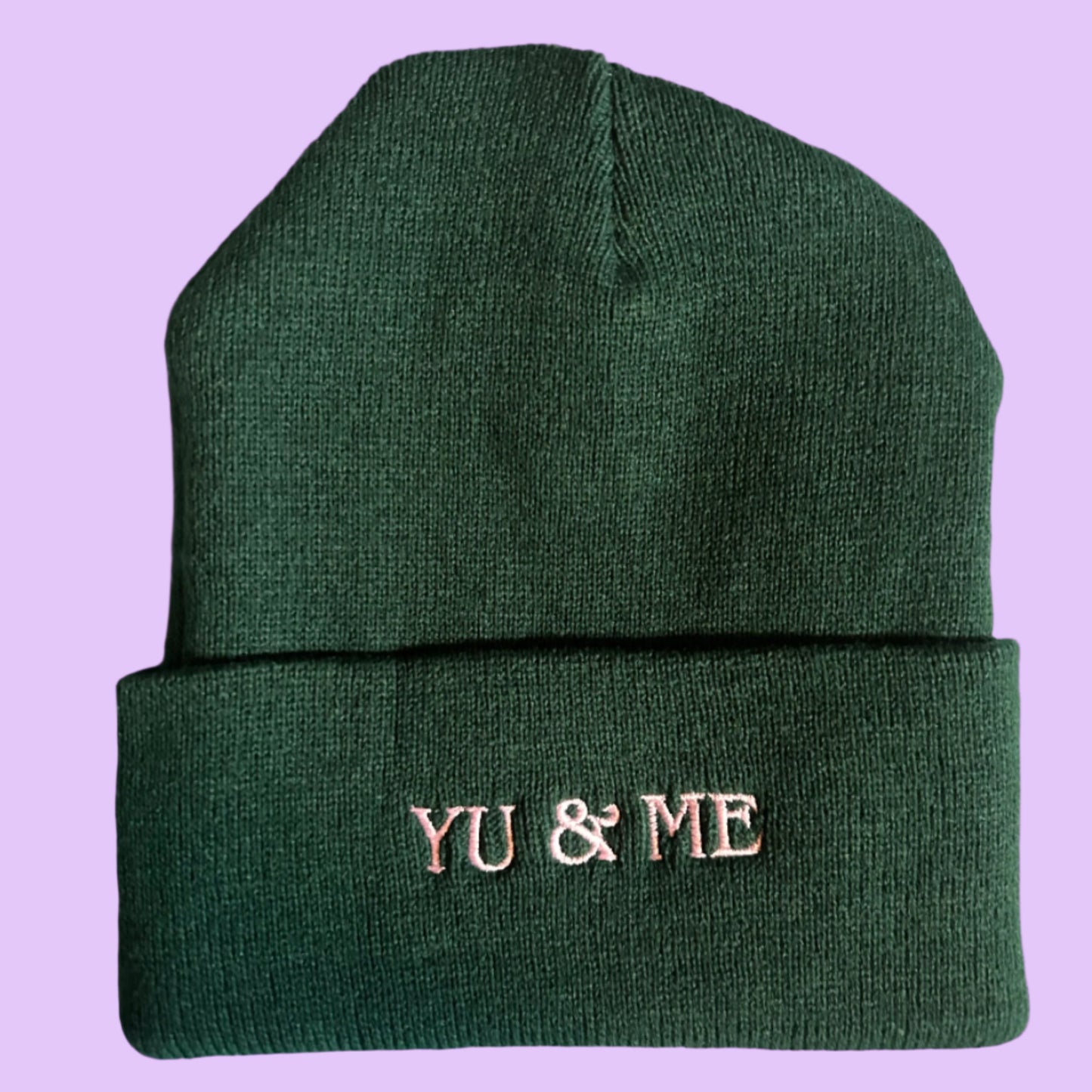 The Yu & Me Beanie by Yu & Me Books is a dark green knit hat with "YU & ME" embroidered in white on the folded brim, shown against a light purple background.