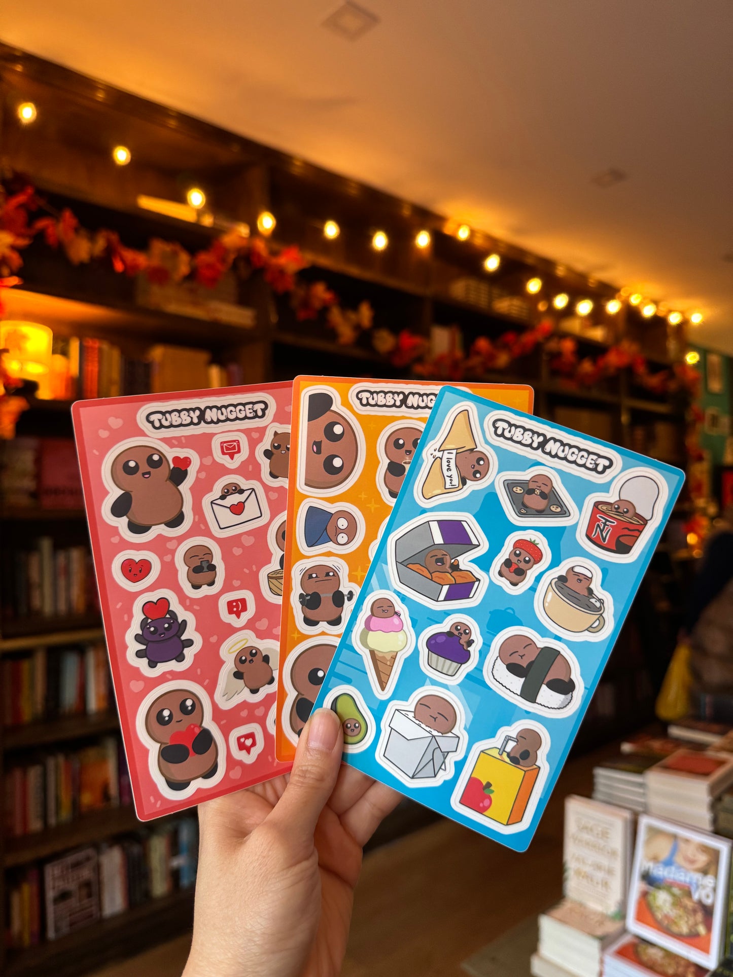 A hand displays three colorful Tubby Nugget sticker sheets in a cozy, warmly lit Yu & Me Books bookstore—perfect to pair with Tubby Nugget's Tearable Book (Signed, with FREE Keychain & Sticker Sheets).