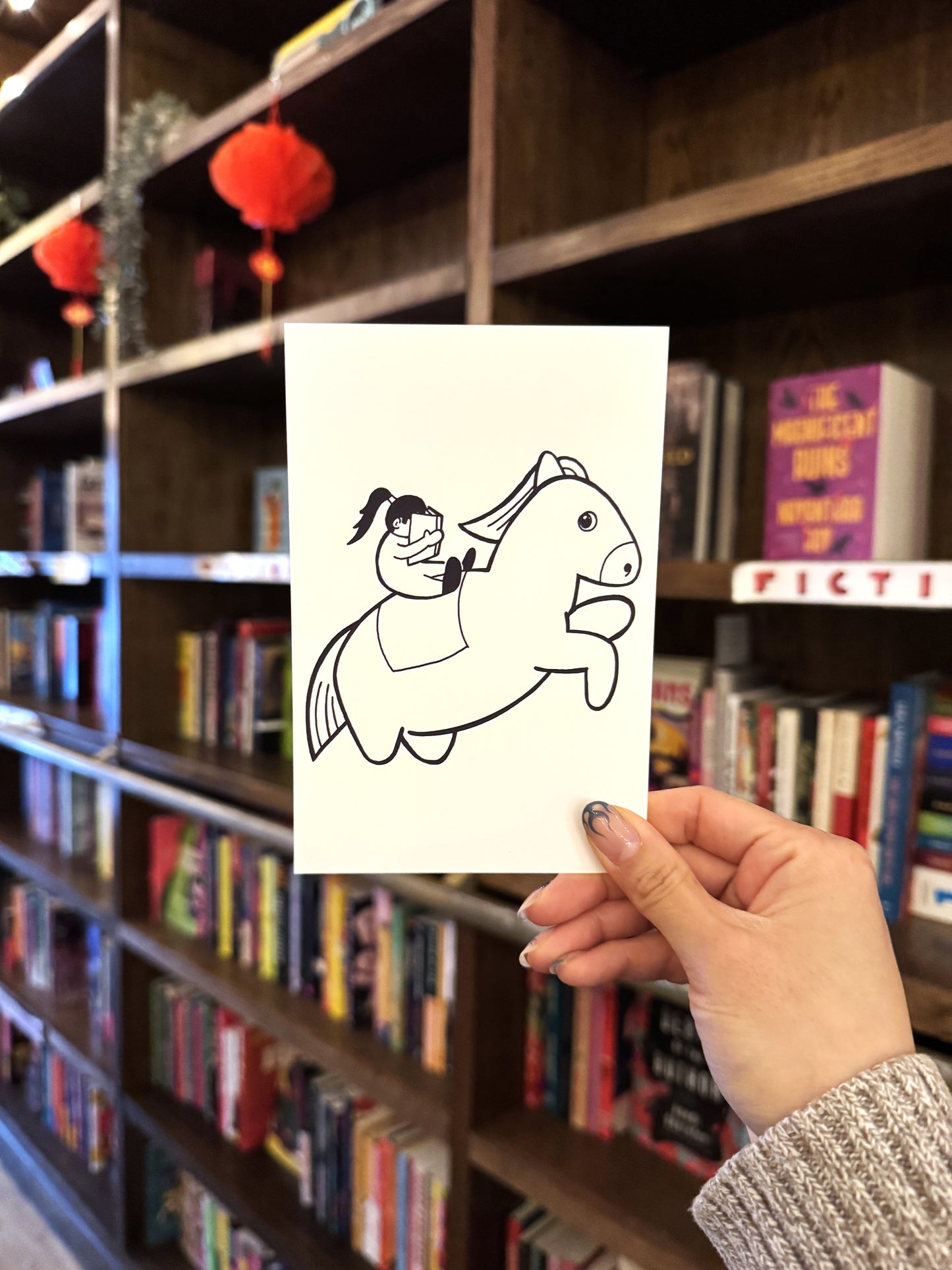 A hand holds the "Year of the (Chonky) Horse, Yu & Me Postcard" from Yu & Me Books, featuring an illustrated girl riding a chonk horse, set against shelves of colorful books and red lanterns.