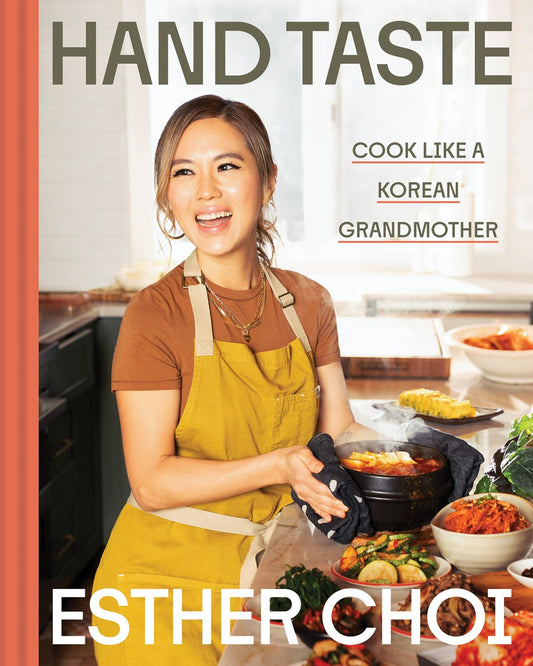 A smiling woman in a yellow apron holds a steaming pot in a bright kitchen with Korean dishes. Nearby is Hand Taste by Esther Choi (Signed & Personalized!) from Yu & Me Books, a cookbook for authentic Korean recipes.
