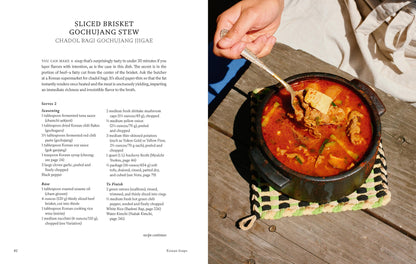 A hand holds a spoon over a black pot filled with vibrant red gochujang stew—generous slices of brisket and tender vegetables peeking through—resting on a patterned mat beside an open copy of Korean Soups by Clara Lee, Eddo Kim (with Signed Bookplate!) from Yu & Me Books, turned to the recipe for sliced brisket gochujang stew.