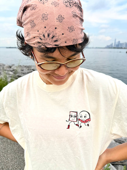 A person in sunglasses, a pink patterned bandana, and the Yu & Me Books 7th Street Burger x Yu & Me Books LIMITED EDITION T-Shirt stands outdoors by the water with a city skyline, celebrating this July exclusive collaboration.