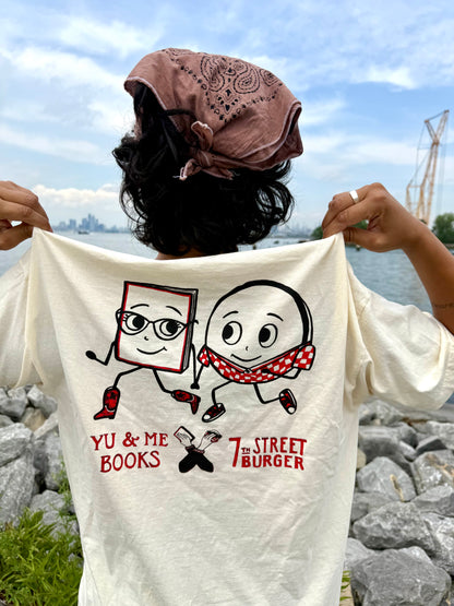 Wearing a pink bandana, a person displays the back of their Yu & Me Books 7th Street Burger LIMITED EDITION T-Shirt—featuring cartoon graphics and collab text—while standing by a rocky lakeshore.