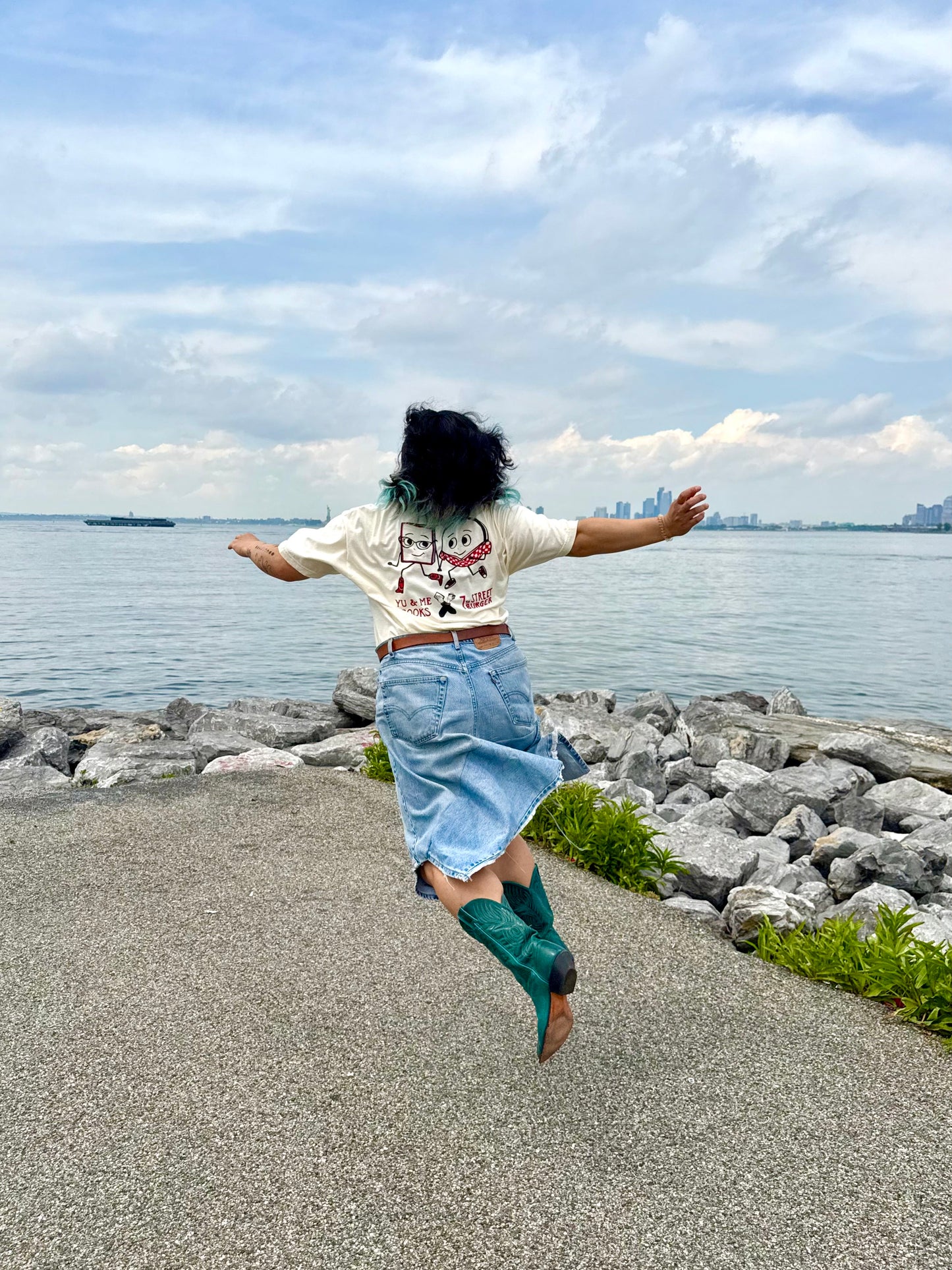 A person with dark hair, wearing the Yu & Me Books 7th Street Burger LIMITED EDITION T-Shirt, denim skirt, and teal boots, jumps joyfully near a rocky lakeshore—celebrating July's exclusive drop with Yu & Me Books.