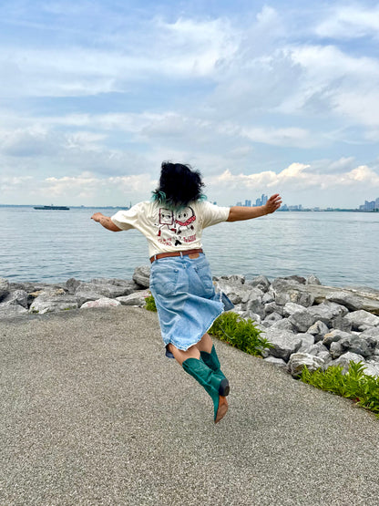 A person with dark hair, wearing the Yu & Me Books 7th Street Burger LIMITED EDITION T-Shirt, denim skirt, and teal boots, jumps joyfully near a rocky lakeshore—celebrating July's exclusive drop with Yu & Me Books.