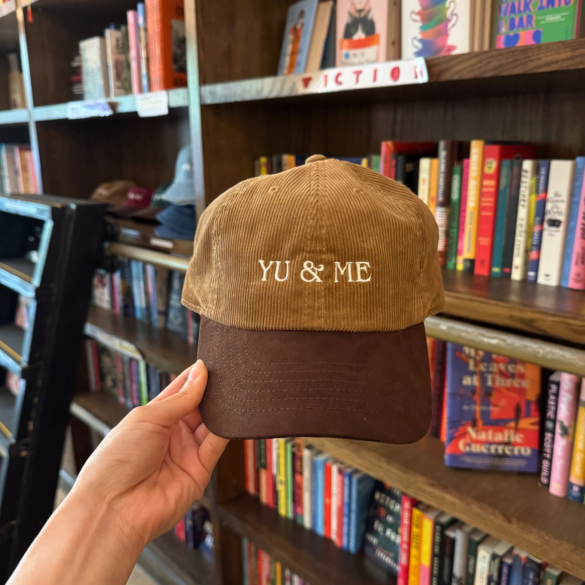 A hand holds the Yu & Me Hat, a brown corduroy baseball cap from Yu & Me Books, in front of bookstore shelves—a cozy scene perfect for fans of stylish hats and good reads.