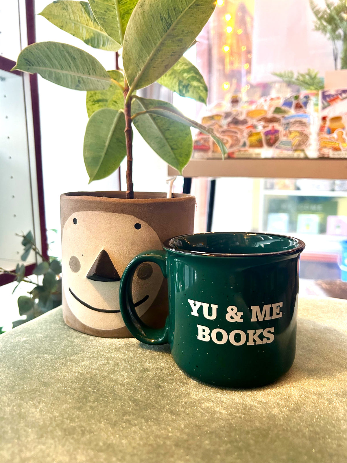 The Green Yu & Me Mug by Yu & Me Books rests on a soft green surface beside a smiley-faced potted plant, with colorful blurred items in the background.