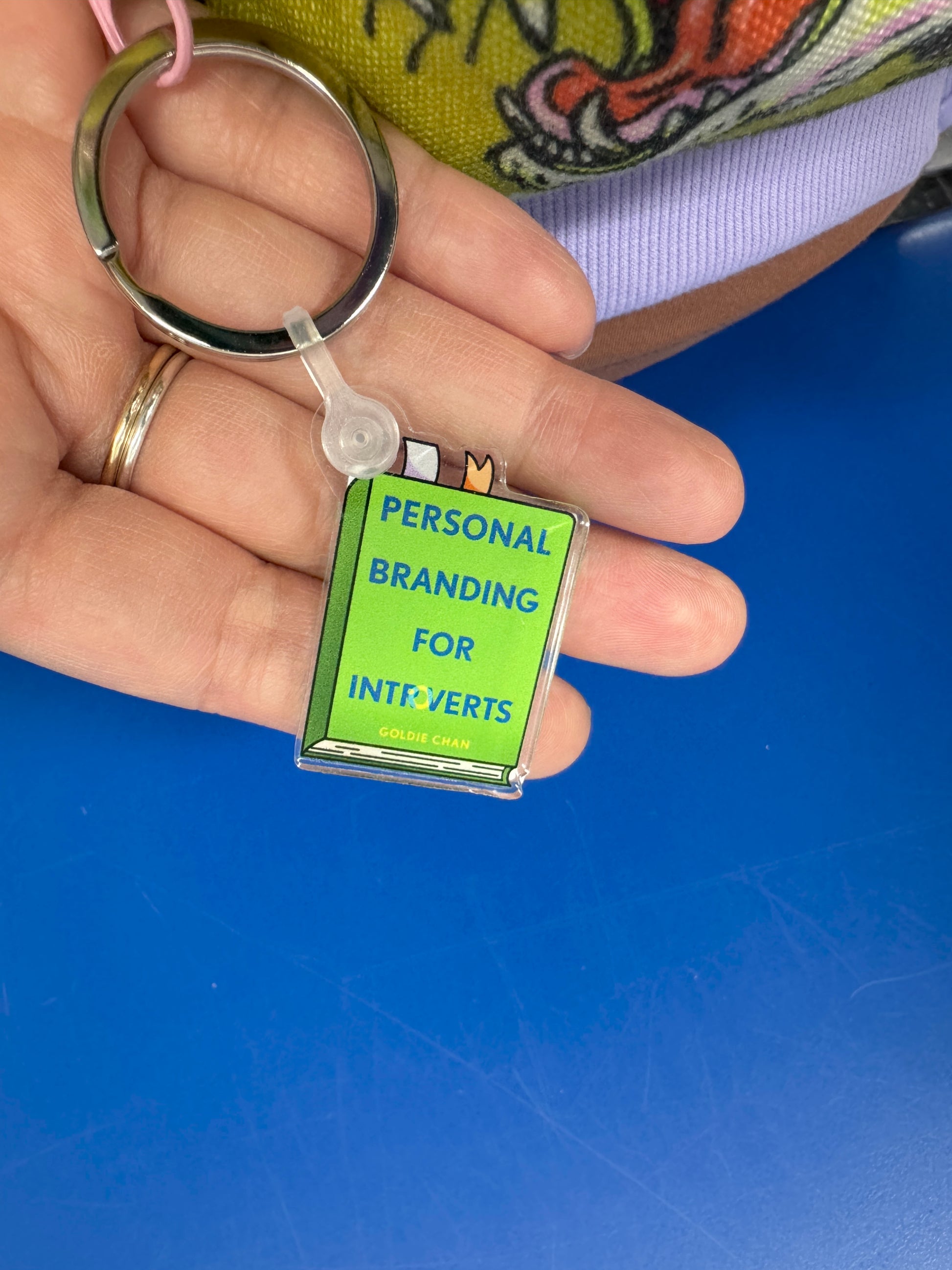 A hand holds a keychain shaped like the green book "Personal Branding for Introverts by Goldie Chan" from Yu & Me Books (signed, personalized, comes with sticker), against a colorful fabric and blue surface background.