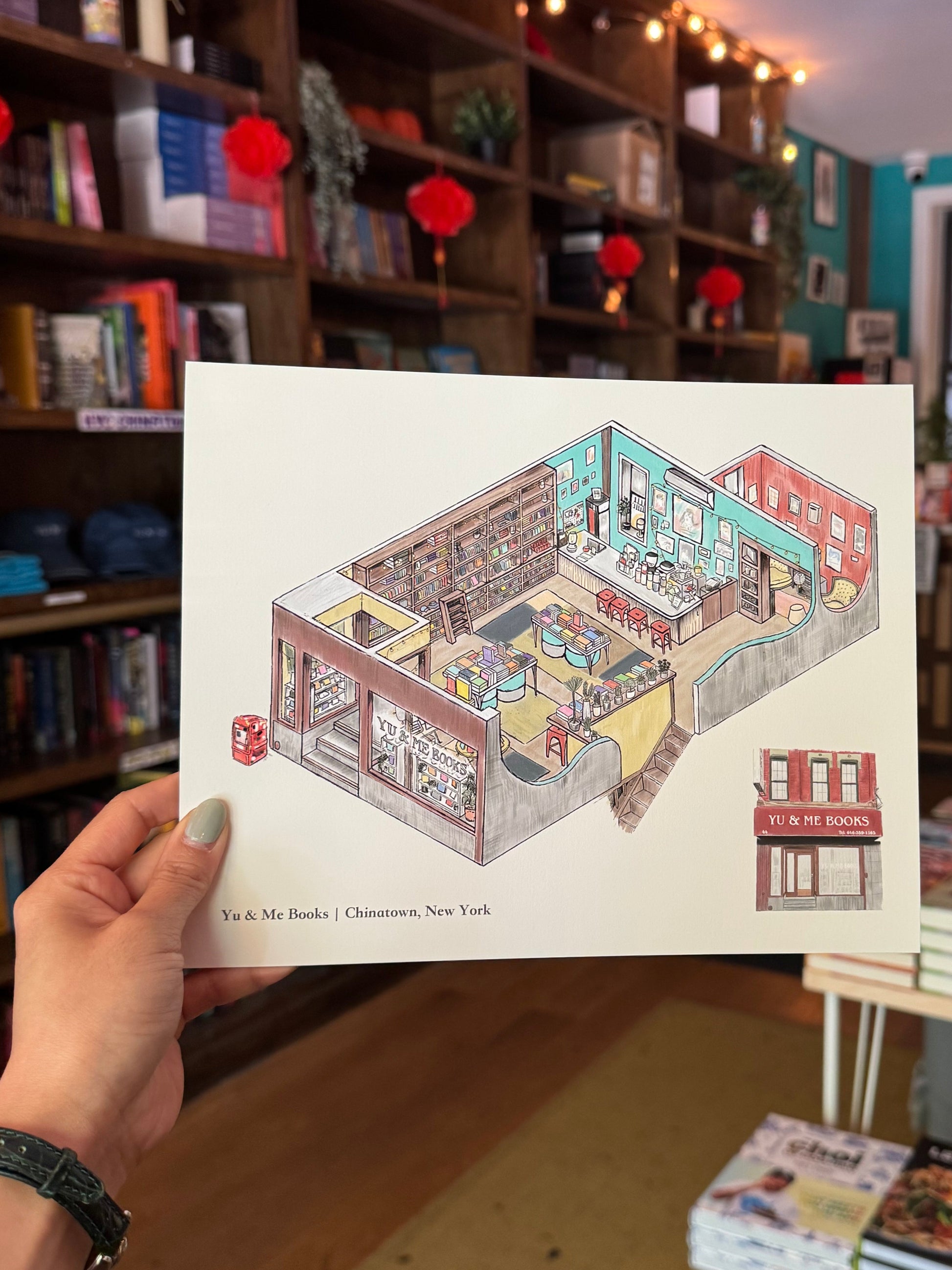 A hand holds the Yu & Me Books "Store Interior" art print, featuring detailed bookshelves and cozy nooks, set at the real Chinatown store—includes a FREE Y&M postcard!.