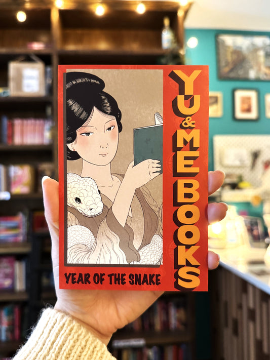 A hand holds the limited-run "Year of the Snake, Yu & Me Post Card" by Yu & Me Books, showing an elegant woman reading with a white snake on her arm against cozy bookshelves and warm lights in the background.