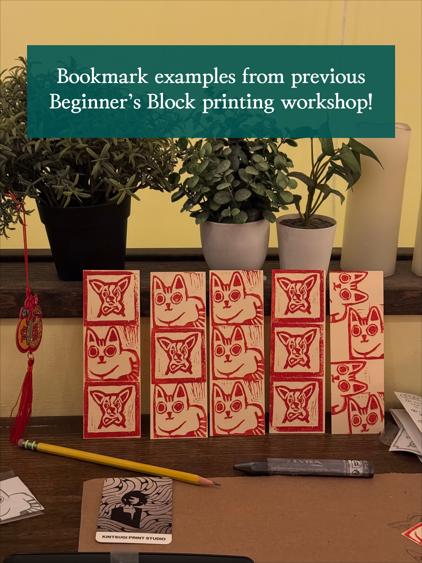 A set of red and white handmade cat and fox bookmarks is arranged on a desk with potted plants. Above, a sign reads: “Bookmark examples from previous Beginner's Block Printing Workshop with Kintsugi Print Studio at Yu & Me Books!”.